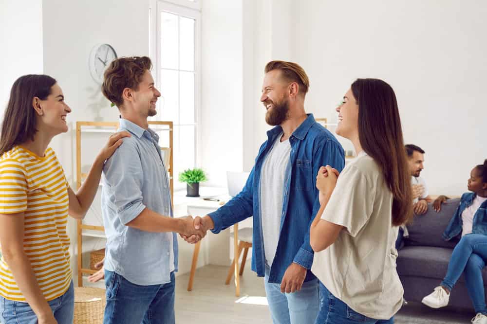 Happy smiling friends greeting and handshaking while meeting. Smiling guy getting acquainted with another at friendly meeting, girls introducing them. Happy people get acquainted hanging together