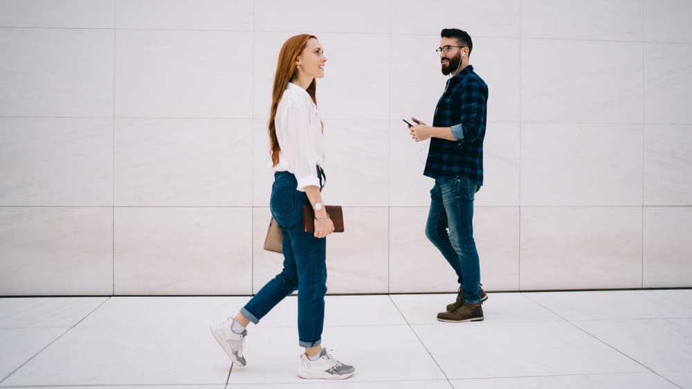 Full body side view of smart casual man and woman passing each other while walking along blank tiled wall on street sidewalk