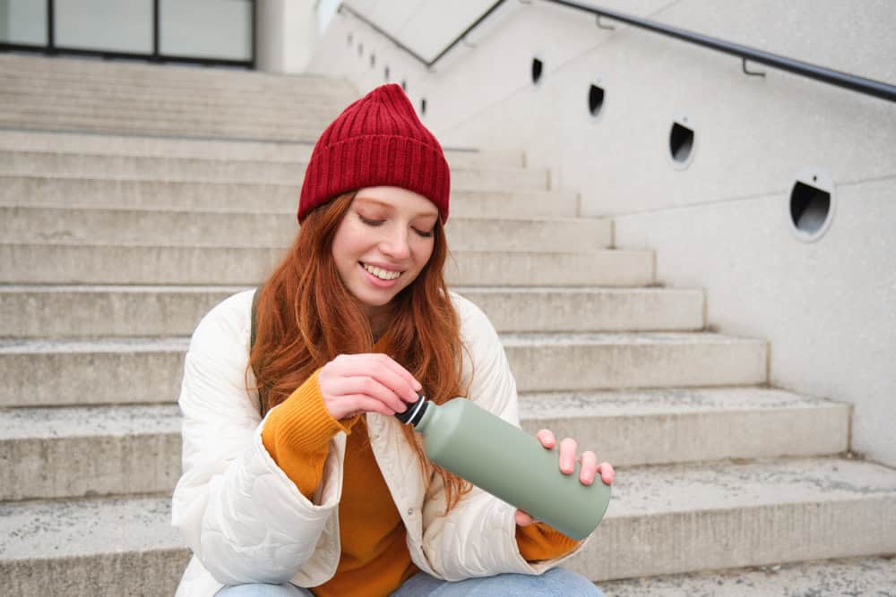 Smiling traveler, redhead girl tourist sits on stairs with flask, drinks hot coffee from thermos while travelling and sightseeing around foreign city, sits on stairs and rests.