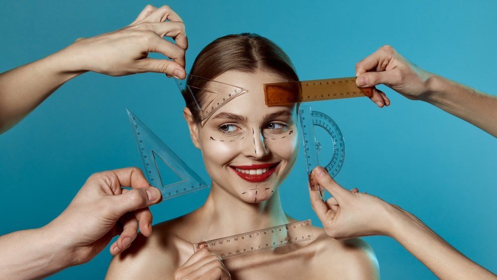 Young happy beautiful woman, female patient with human hands holding ruler isolated on blue studio background. Lifting concept. Correction of asymmetry.