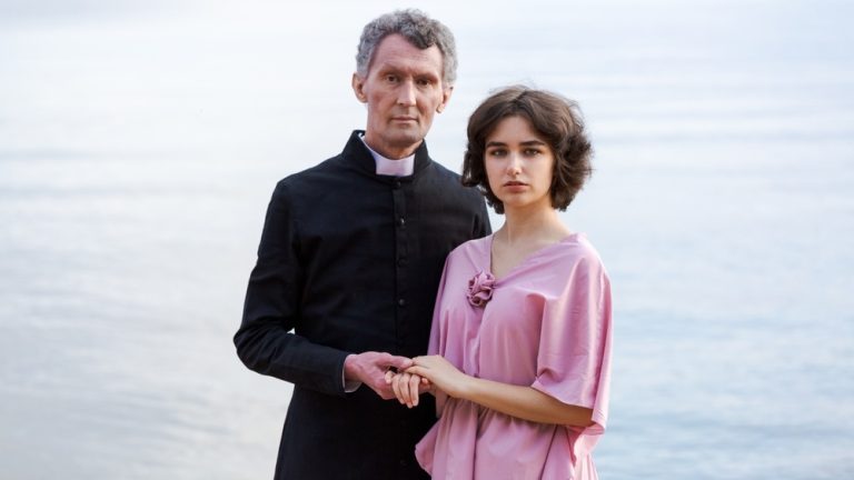 a young woman in a pink dress and an elderly man stand together on the seashore at sunset. the concept of the relationship of different ages in a couple