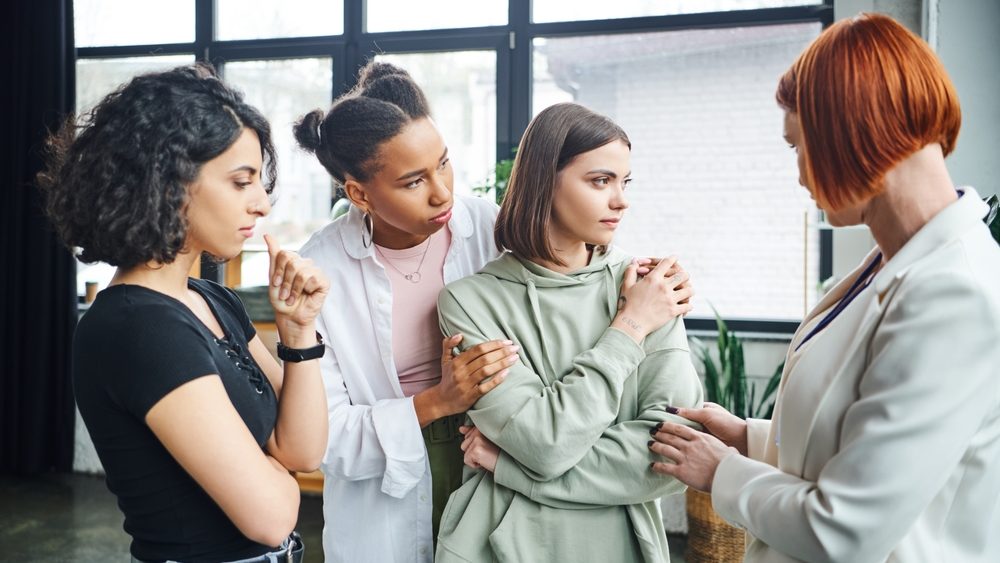 multiethnic woman calming young frustrated girlfriend near motivation coach during psychology session in consulting room, problem-solving and psychological help concept