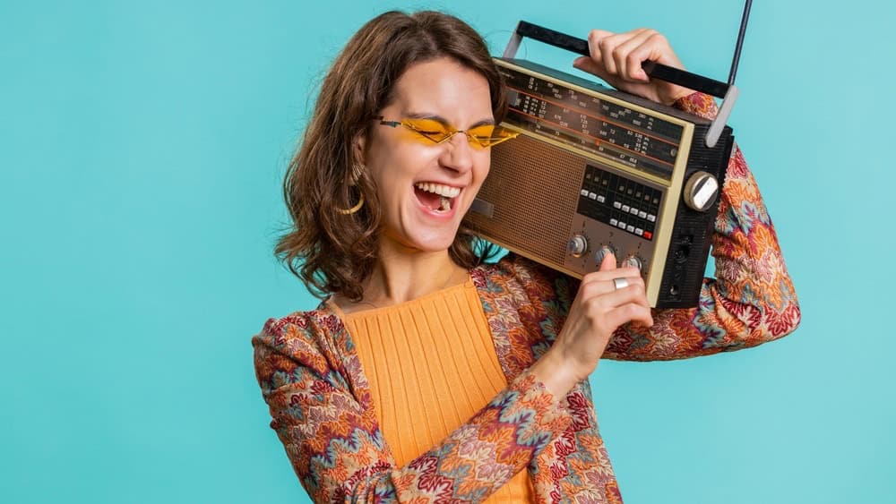 Woman in sunglasses using retro tape record player to listen music, disco dancing of favorite track, having fun, entertaining, fan of vintage technologies. Brunette girl isolated on blue background