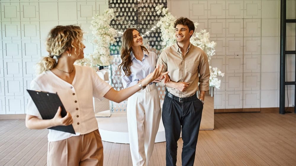 professional event manager with clipboard showing wedding venue with floral decor to happy couple