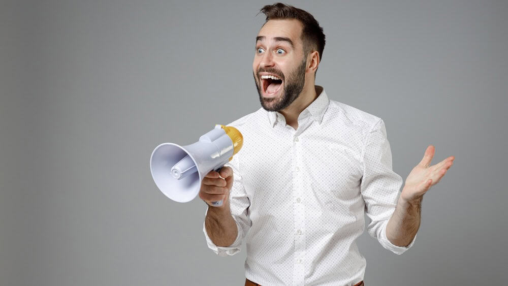 Surprised young bearded business man 20s in classic white shirt standing screaming in megaphone spreading hands isolated on grey color background studio portrait. Achievement career wealth concept