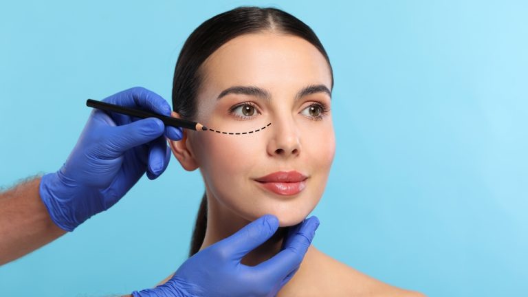 Woman preparing for cosmetic surgery, light blue background. Doctor drawing markings on her face, closeup
