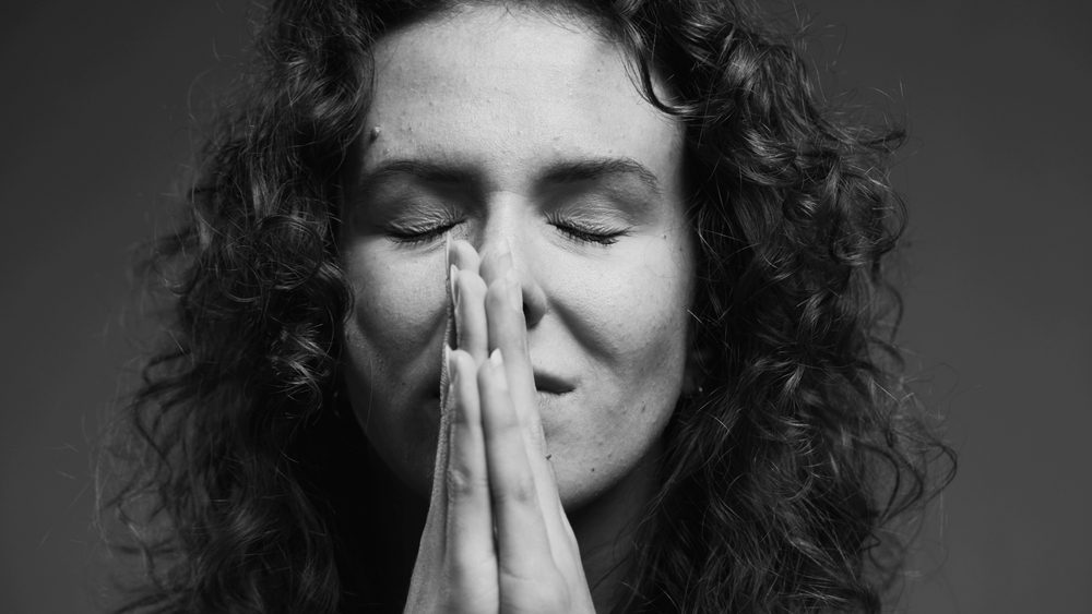 Spiritual 20s person feeling GRATITUDE and Godly devotion with eyes closed and hands clenched in PRAYER. Hopeful woman having FAITH in monochromatic, black and white