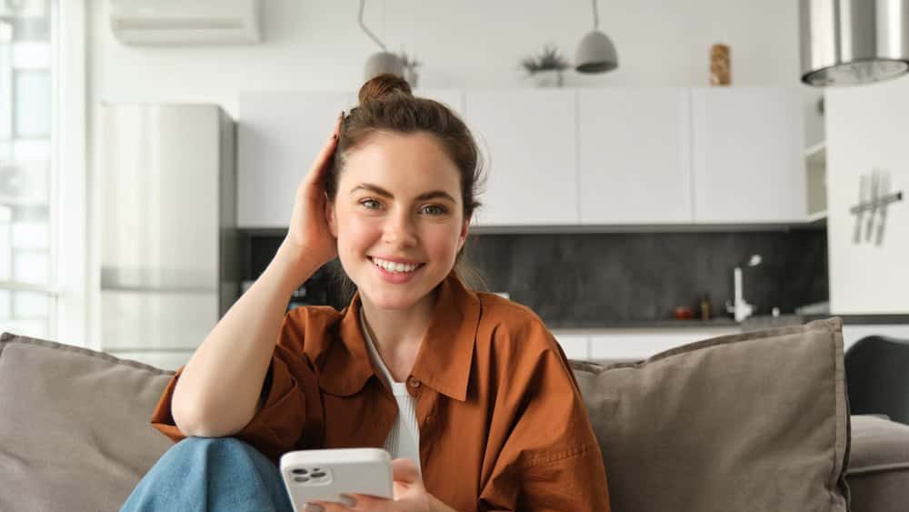 Beautiful smiling female model sitting on couch with smartphone, looking happy at camera, scrolling social media, messaging someone, doing online shopping.