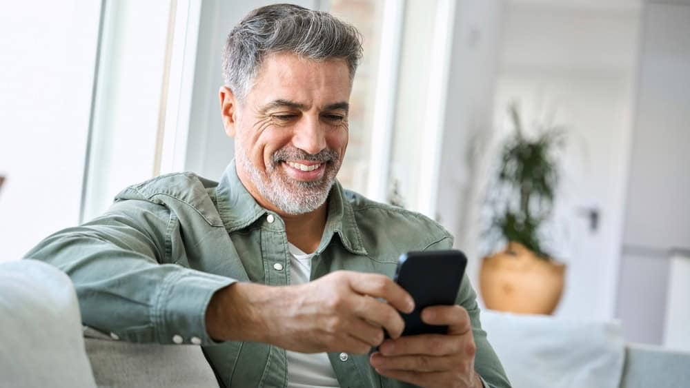 Smiling happy senior mature middle aged man holding cell mobile phone using smartphone sitting at home on couch, scrolling social media, checking financial apps, buying online, texting messages.