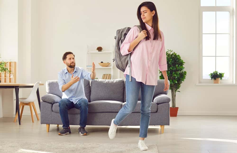 Young wife with packed backpack leaving her husband sitting on sofa at home. Unhappy couple man and woman breaking up after quarrel. Divorce, relationship and marriage problems concept.