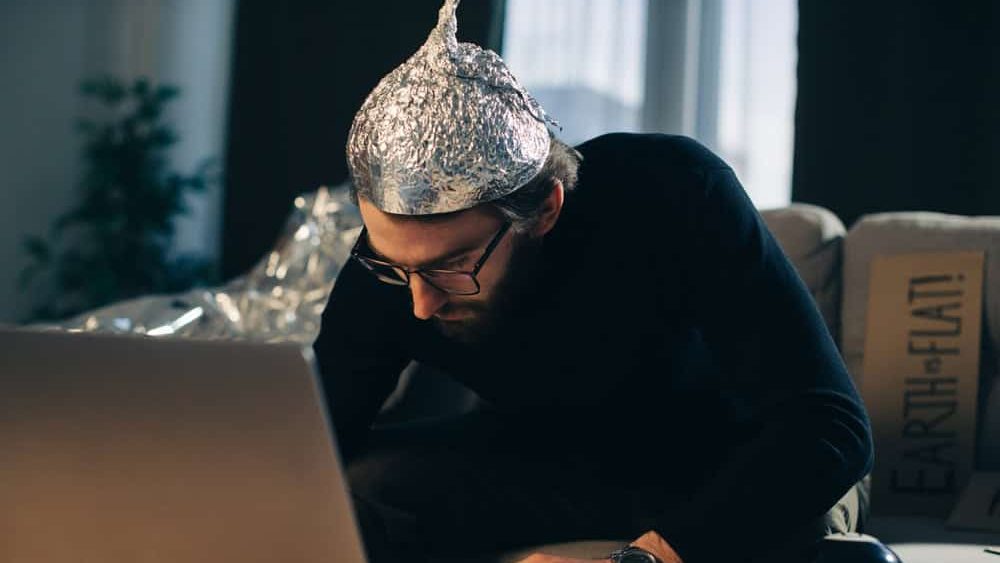 Conspiracy theory. A man in a tinfoil hat looks for signs while watching a video on a laptop.