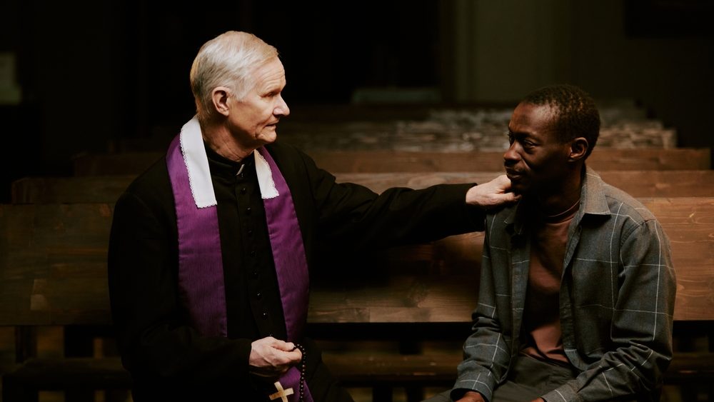 Medium shot of senior Caucasian Catholic priest and young African American man sitting on pew in church having conversation