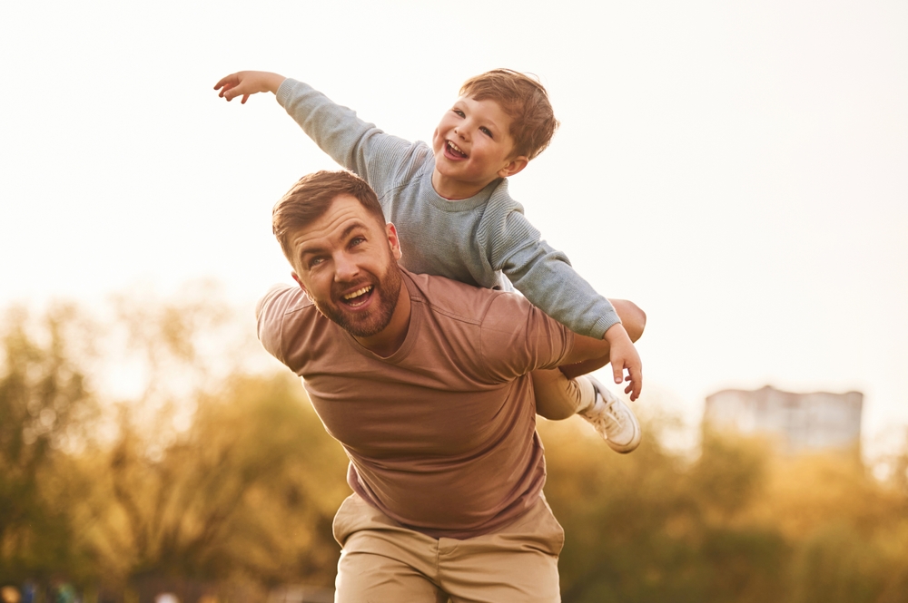 Conception of imagination, sitting on shoulders and flying. Happy father with son are having fun on the field at summertime.