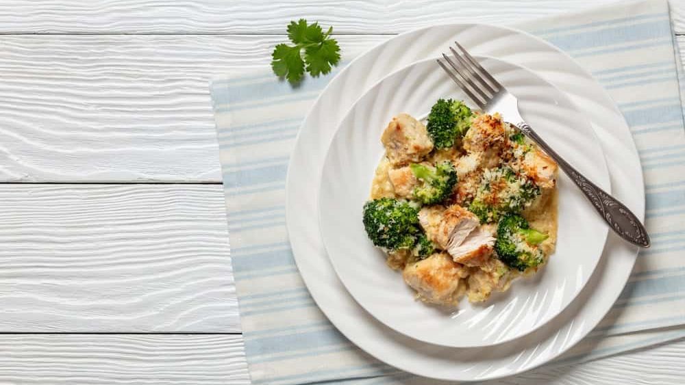 portion of Chicken Divan, succulent chicken bites in creamy sauce with broccoli topped with panko on white plate with fork on white wooden table, american cuisine, flat lay, free space
