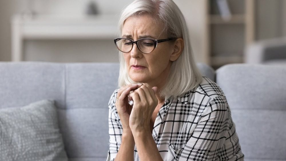 Upset stressed mature woman in glasses sitting alone on couch at home, suffering from anxiety or incurable disease, feeling loneliness, desperation, looks frustrated, mourning, grieving about bad news