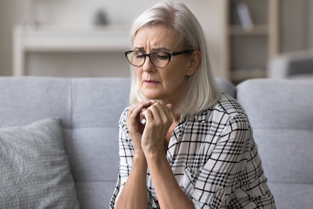 Upset stressed mature woman in glasses sitting alone on couch at home, suffering from anxiety or incurable disease, feeling loneliness, desperation, looks frustrated, mourning, grieving about bad news