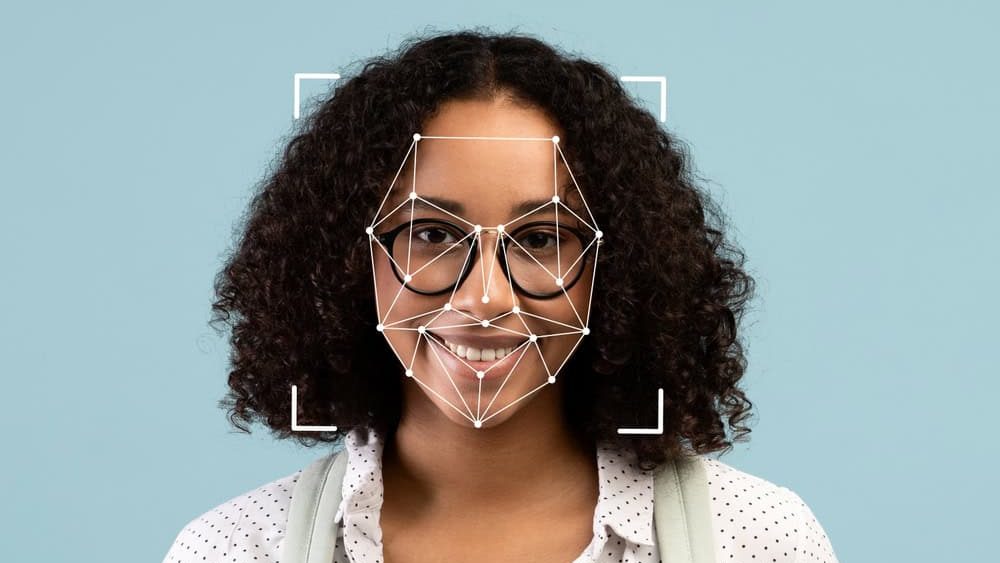 A young African American woman with curly hair and glasses smiles at the camera, showcasing a facial recognition overlay against a soothing blue background.