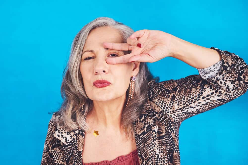 Peace sign, dance and portrait of senior woman in studio for fun, comedy and funky style at party. Confident person, fashion and gesture with v on eye for retirement celebration by blue background