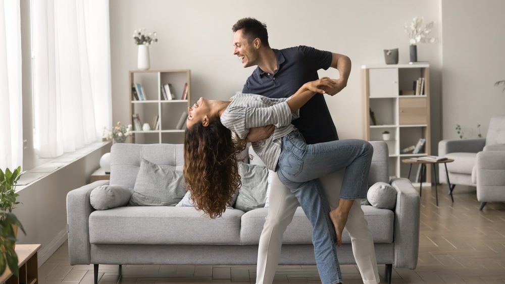 Happy active married couple dancing in stylish living room interior, enjoying activity at home together. Positive strong husband embracing wife in romantic social dance with passion