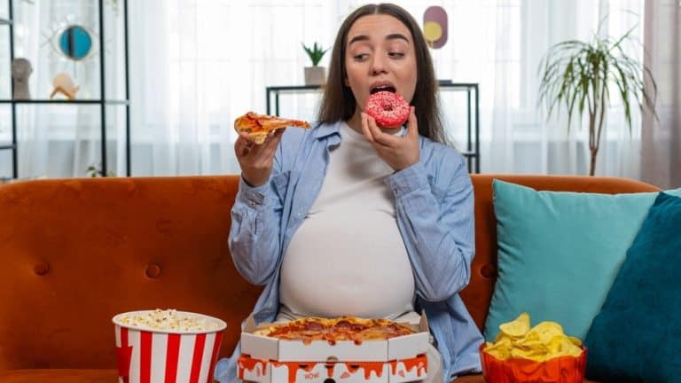 Pregnant woman eating unhealthy junk food pizza, popcorn crisp sitting on sofa in home living room. Fast fatty food during pregnancy in apartment. Hunger, change in taste preferences. Expecting baby