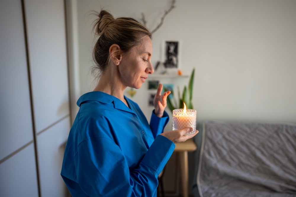 Calm middle-aged woman enjoy smelling lighted scented aromatic candle in crystal, meditating, filling home with pleasant smell, creating cozy atmosphere. Coziness, healthy habit, mindfulness lifestyle