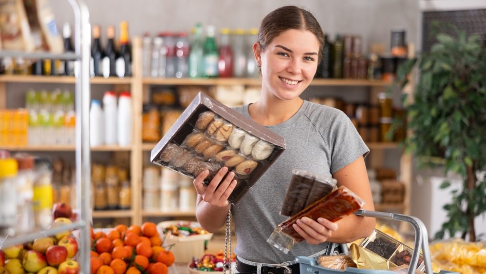 Young caucasian woman chooses sweet desserts in a supermarket with a fruit stand in the background. Customer chooses shortbread and oatmeal cookies in the pastry section
