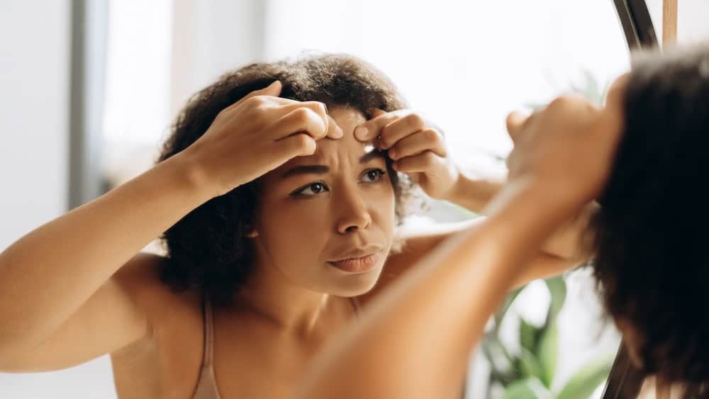 Young woman examining her reflection in the mirror while squeezing a pimple on her forehead, focusing on her skincare routine in the bathroom