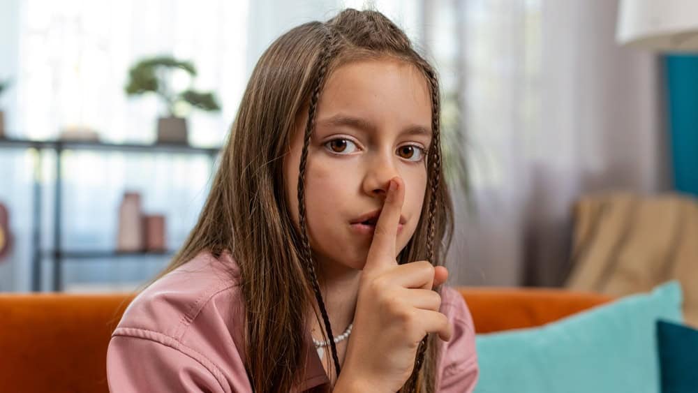 Shh be quiet please. Portrait of preteen girl child presses index finger to lips makes silence hush gesture sign do not tells gossip secret. Little cute teenage female sitting on sofa in living room.