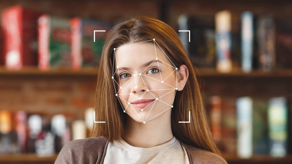 A young woman smiles as facial recognition technology analyzes her features in a library surrounded by books. The focus is on her face, highlighting advanced ID technology in use.