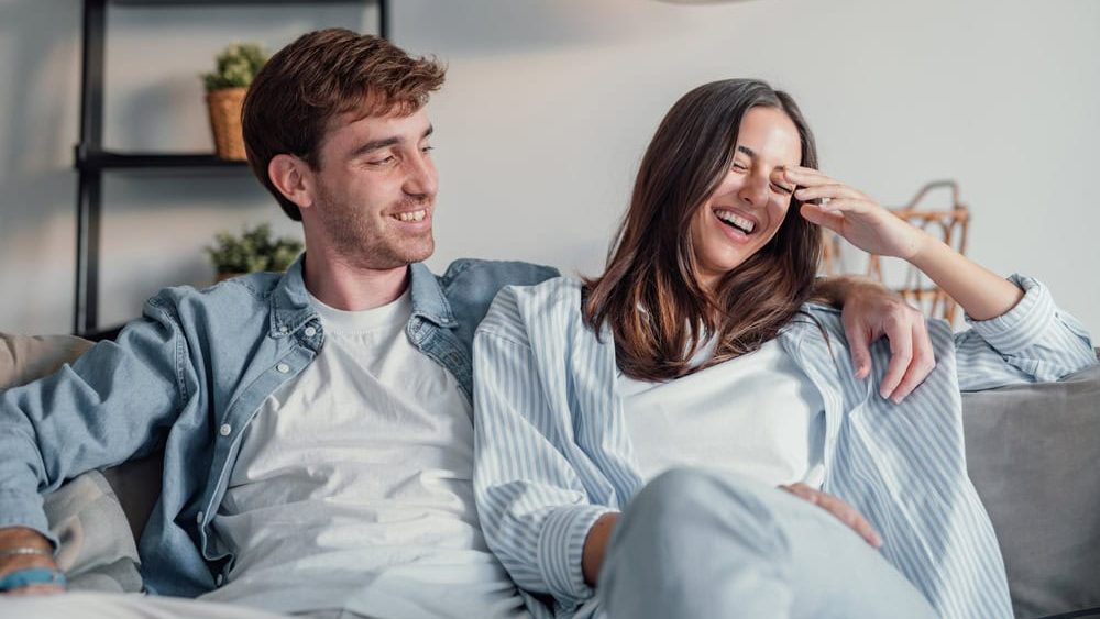 Young happy couple having fun talking laughing relaxing at home on couch, boyfriend embracing girlfriend telling funny joke sitting on sofa, humor in relationships, enjoying weekend together
