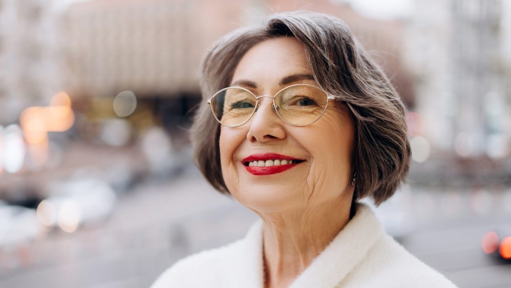 Elegant senior woman with short gray hair and glasses, smiling confidently in an urban environment. Wearing a white coat she exudes warmth and positivity amid a bustling city backdrop. Fashion concept