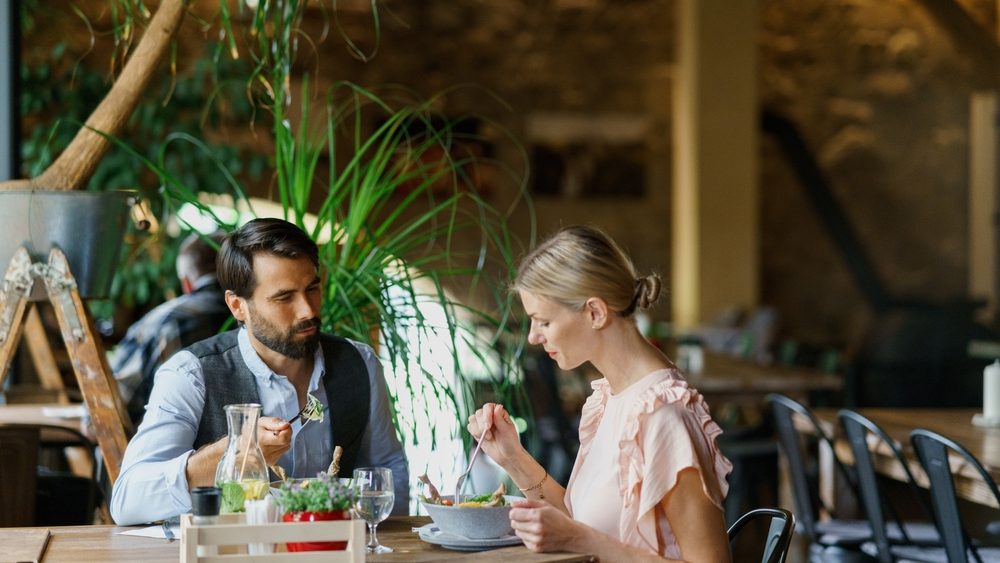 Happy loving couple at date in restaurant. Husband and wife sitting at table, talking, having lunch in modern cafe.