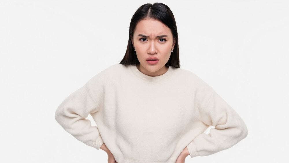 Portrait of displeased offended young Asian woman with black hair keeps arms folded looks angrily aside doesn't agree with somebody opinion wears casual clothes isolated over white studio background