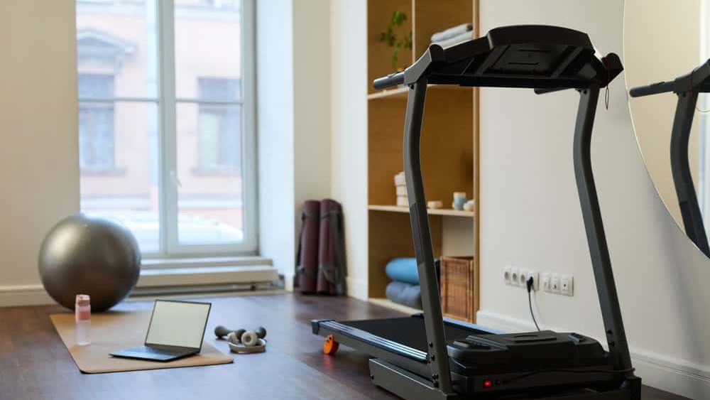Cozy home gym with treadmill positioned in front of a window, exercise ball, laptop and water bottle encouraging a workout routine