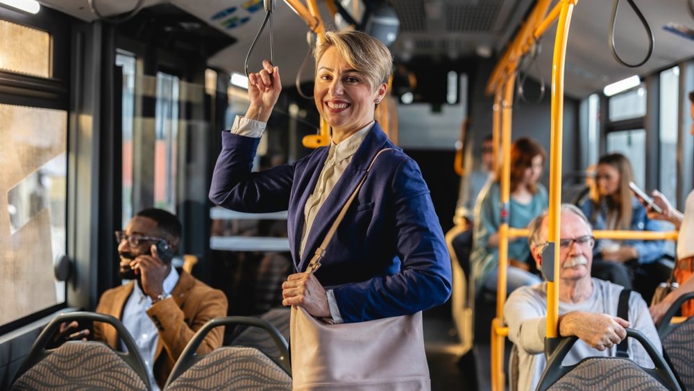 Cheerful businesswoman commuting and holding a handrail while standing on a moving bus, other people sitting and standing in the background