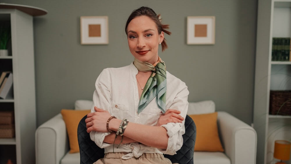 Businesswoman wearing a silk scarf and bracelets sits with folded arms at her desk in a stylish home office, exuding confidence and professionalism