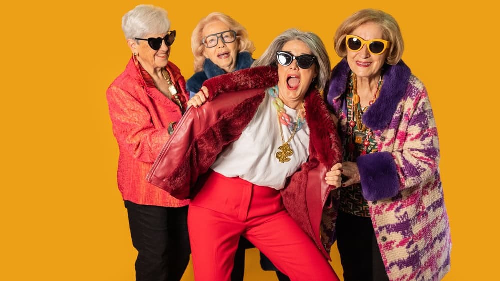 Four fashionable senior women are striking a pose, expressing their unique style with colorful outfits, sunglasses, and playful attitudes against a vibrant yellow backdrop