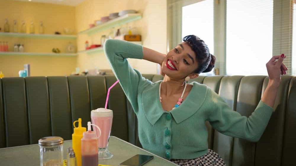 Smiling woman stretching arms behind head at diner booth, with milkshake and smartphone, copy space. Vintage, retro, pastel, café, nostalgic, lifestyle, cheerful