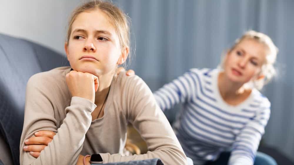 Upset teenager girl after quarrel with her mother in domestic interior, mother consoles daughter