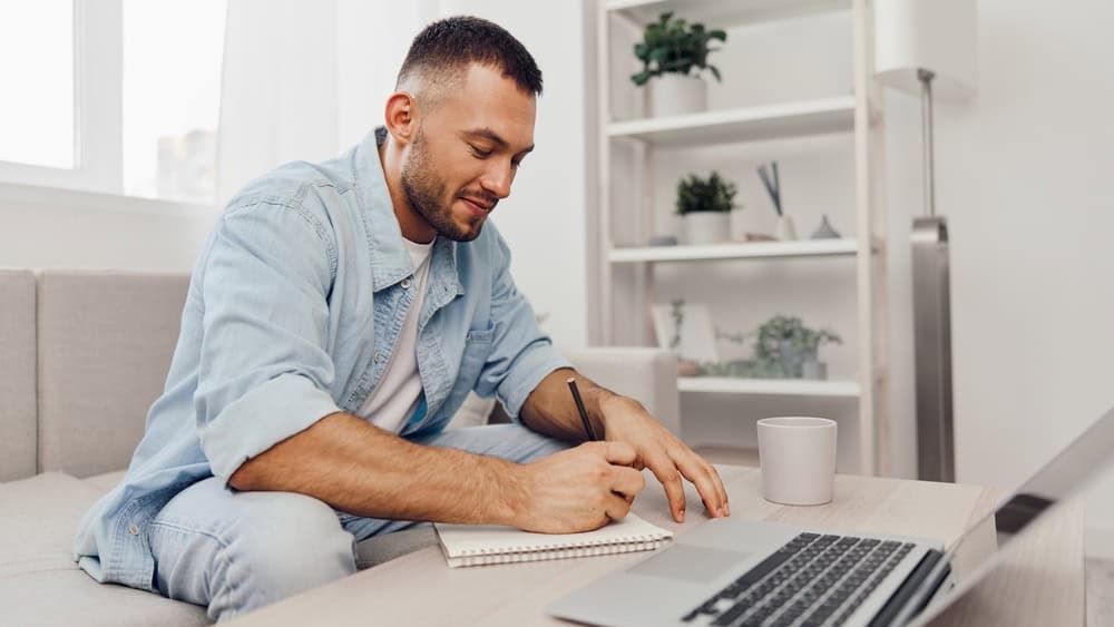 Young man writing in a notebook while sitting on a couch, showcasing creativity and productivity in a cozy, modern workspace environment.