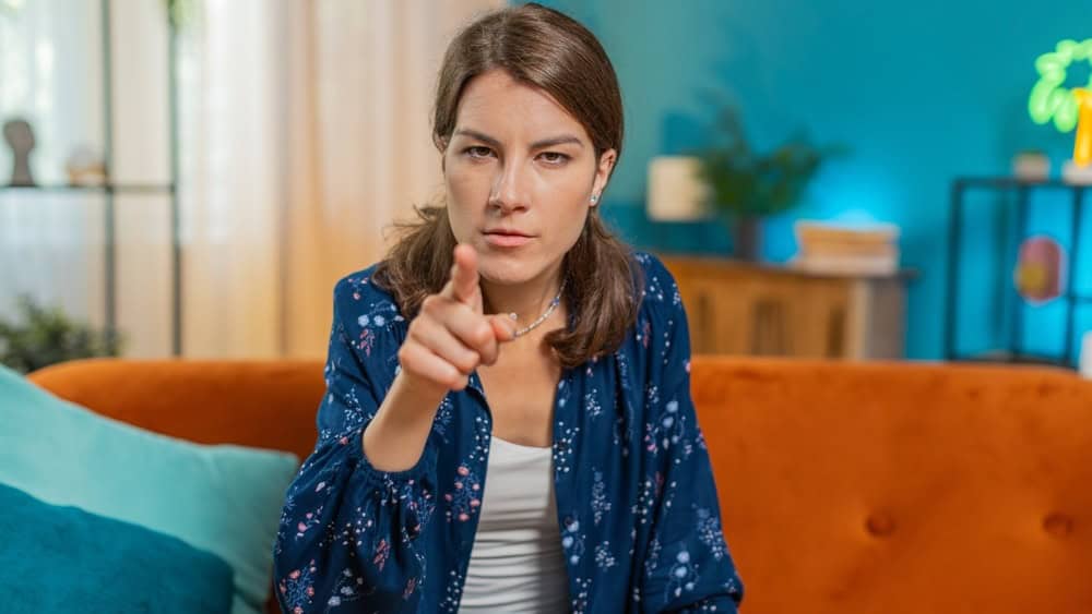 Furious young Caucasian woman sitting on sofa couch at home, angrily pointing at you through the camera, displeased and fuming. Young adult girl in serious blame mode, upset, and ready to explode.