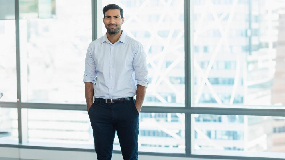 Asian man standing with hands in pockets in high-rise office with glass windows overlooking skyline. Professional, corporate, modern, sleek, urban, workspace, confident