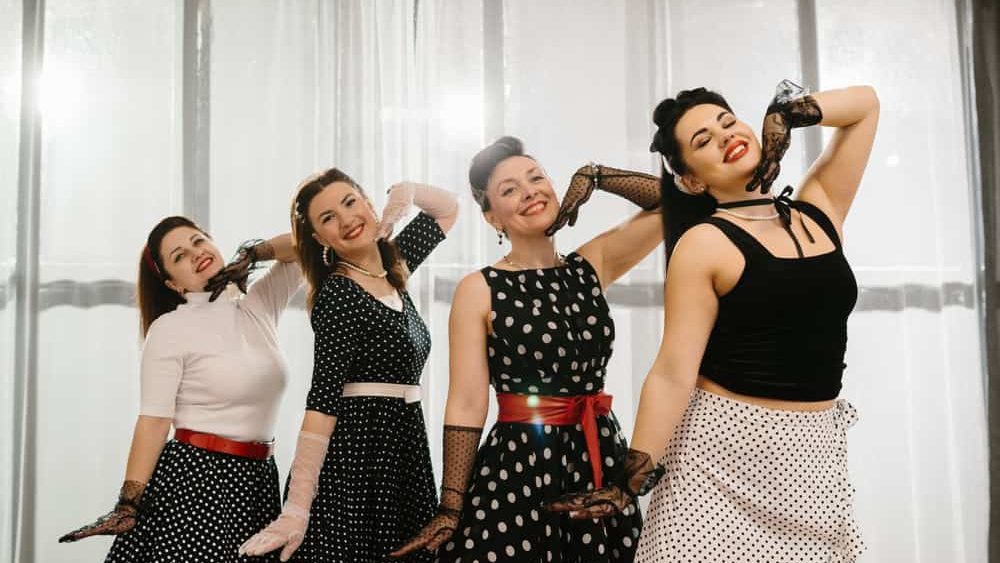 Four women wearing 1950s style polka dot dresses and gloves are posing and dancing in a bright room