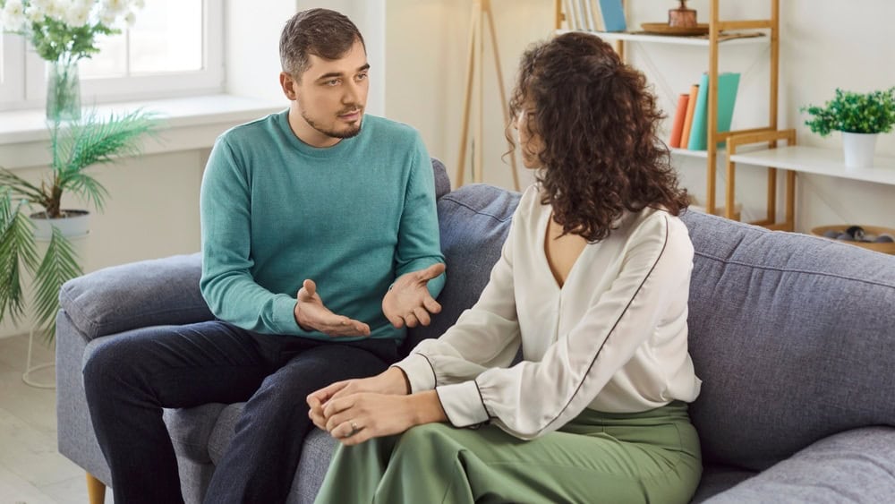Annoyed man sitting on sofa at home and arguing with his wife. Stressed family couple having tense conversation, expressing anger and disappointment during conversation. Relationship problems concept.