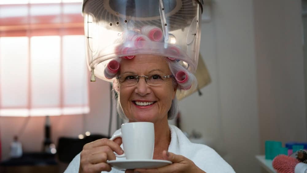 Senior woman smiling under dryer hood with pink rollers at styling station holding mug and saucer. Beauty, wellness, relaxation, elegance, skincare, self-care, comfort