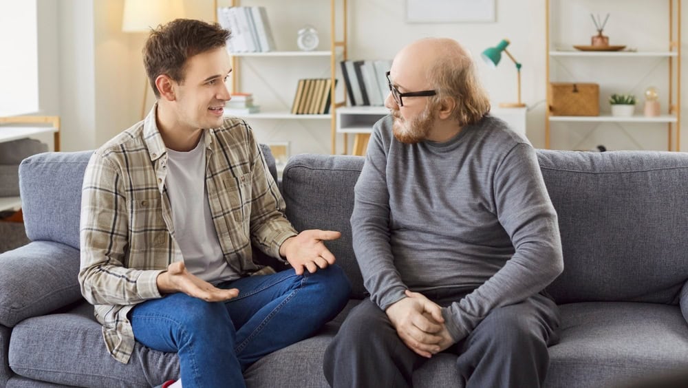 Adult son and a senior father talking. Old man in his 60s sitting on the couch together with his grown up son and listening to him ready to share his life wisdom. Parents and children concept