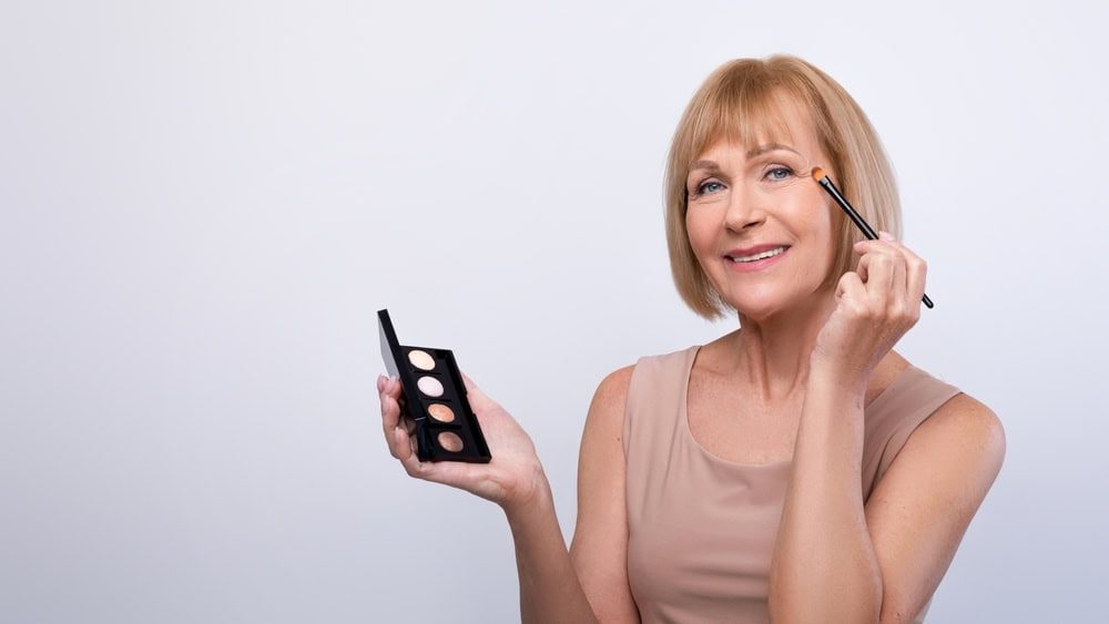 Beauty portrait of senior woman applying eye shadow with cosmetic brush, using swatches palette on light background