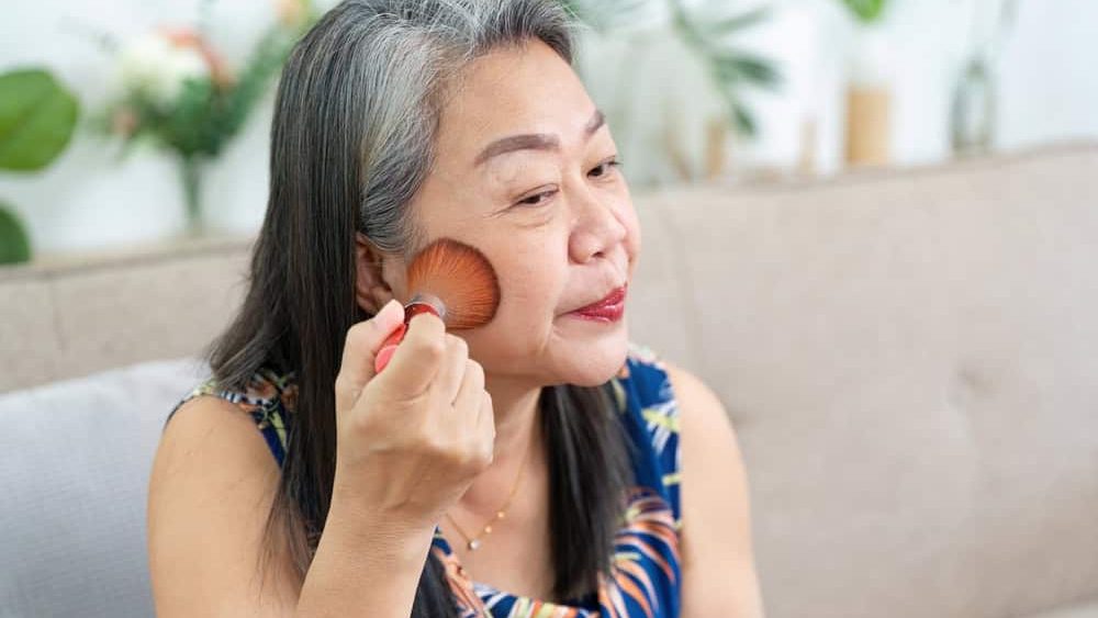 Makeup Application. Mature woman applying blush with a brush in front of a mirror.