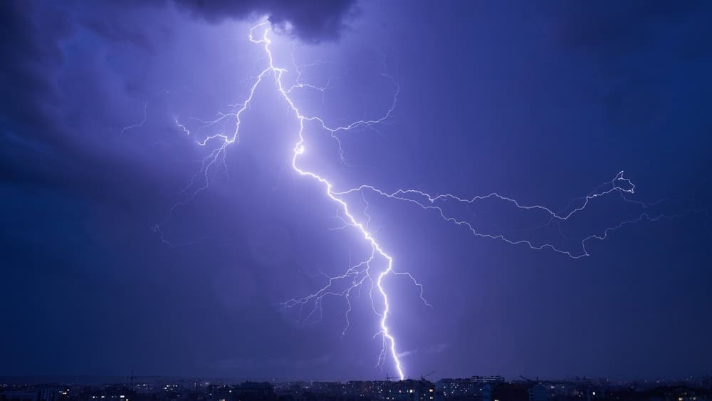 Powerful bolt of lightning cutting through dark, stormy sky over cityscape at night. Lightning's bright illuminate scene, contrasting sharply with dark clouds and dim lights of buildings below.