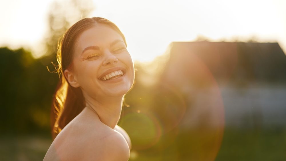 Happy young woman laughing in sunlight, wearing a white off shoulder dress, with a warm golden background, conveying joy and positivity in a summer setting